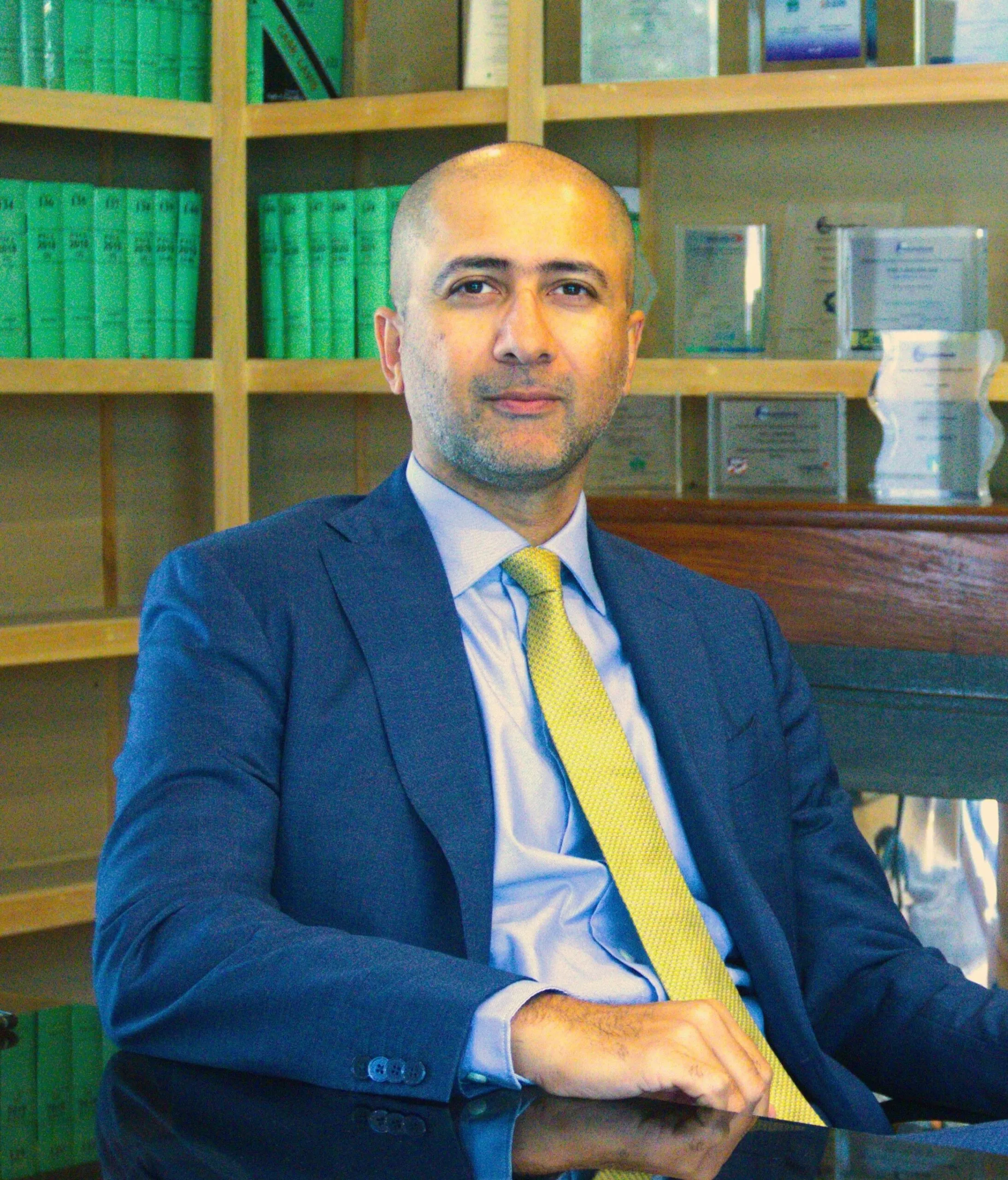 Lawyer seated at office desk with legal books and awards displayed in the background.