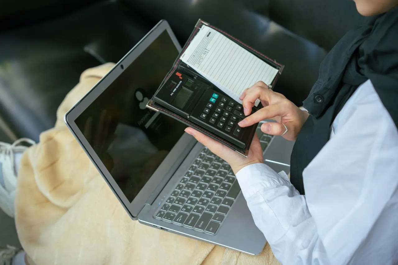 Legal professional using a calculator while reviewing notes next to a laptop at a workspace.