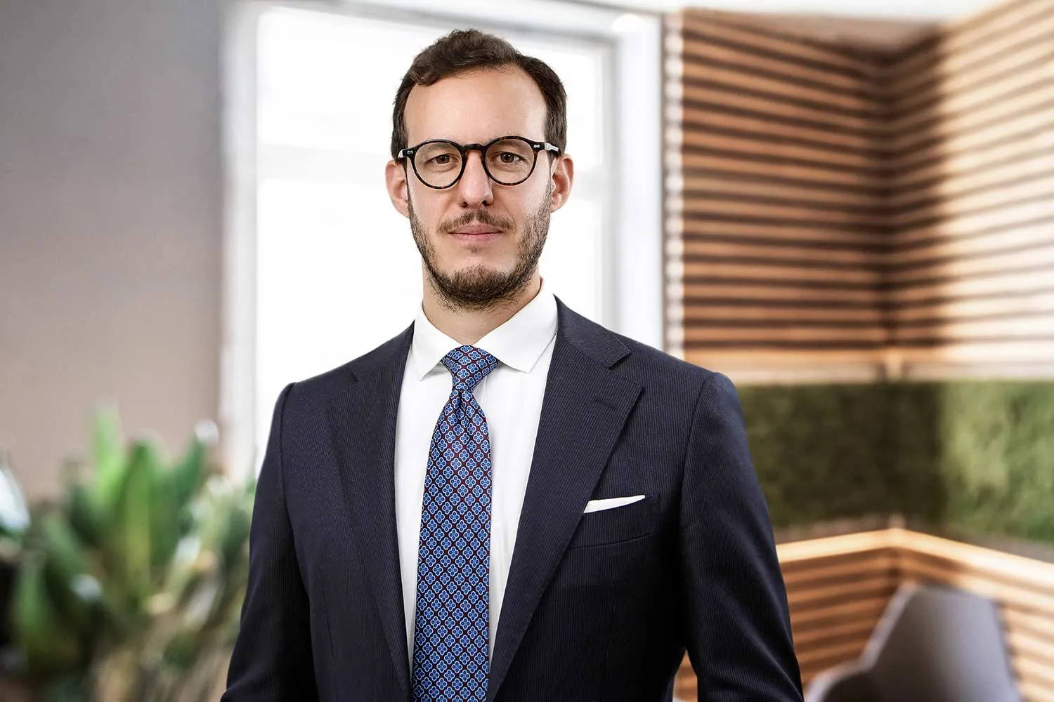 Attorney in a suit standing confidently in a modern office setting with natural light and green plants in the background.