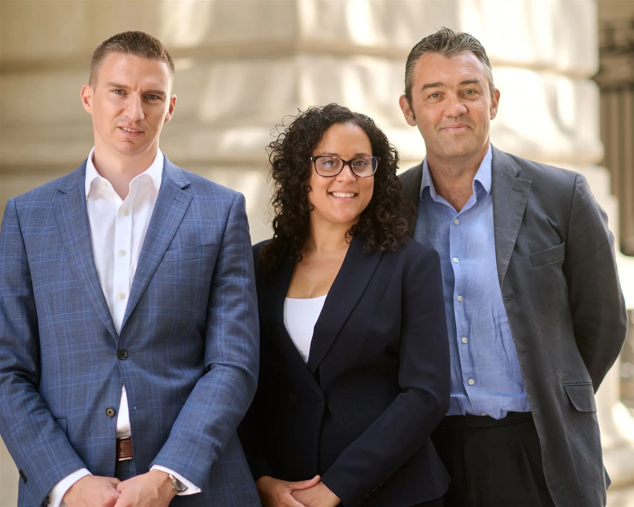 Three legal professionals posing together outside a building, wearing business attire and smiling at the camera.