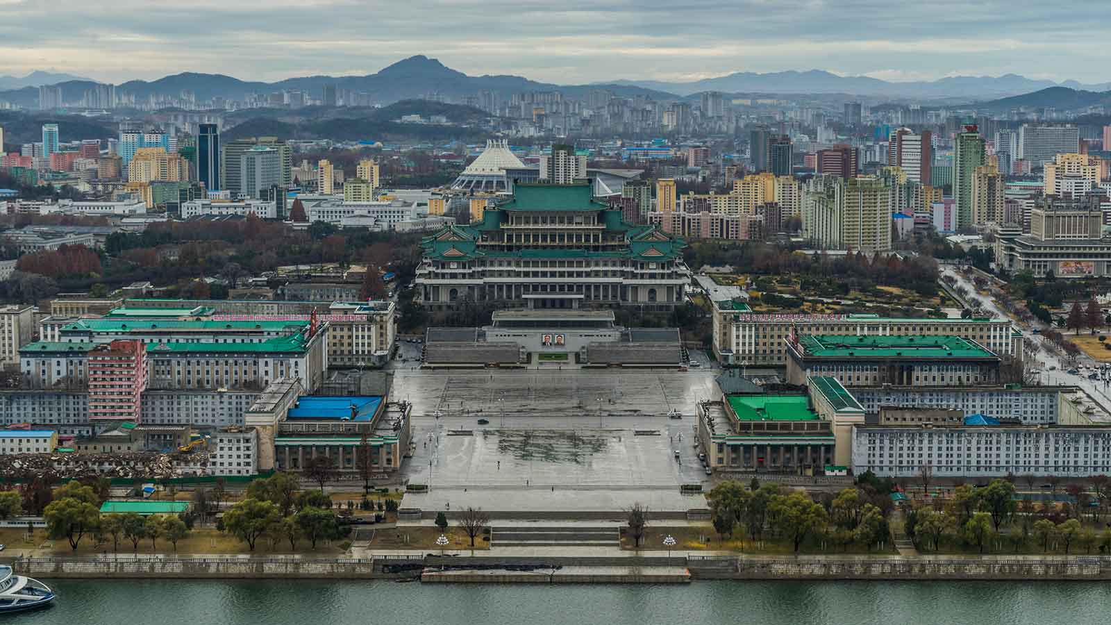 Cityscape featuring a governmental building and modern architecture along a riverbank with mountains in the background.