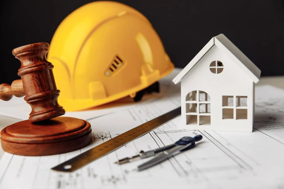 Gavel placed on blueprints with a white model house, yellow hard hat, ruler, and compass on a work surface.