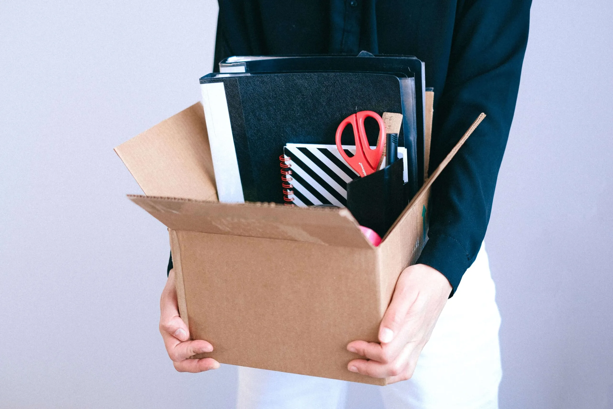 Individual holding a cardboard box filled with office supplies, including folders and scissors.