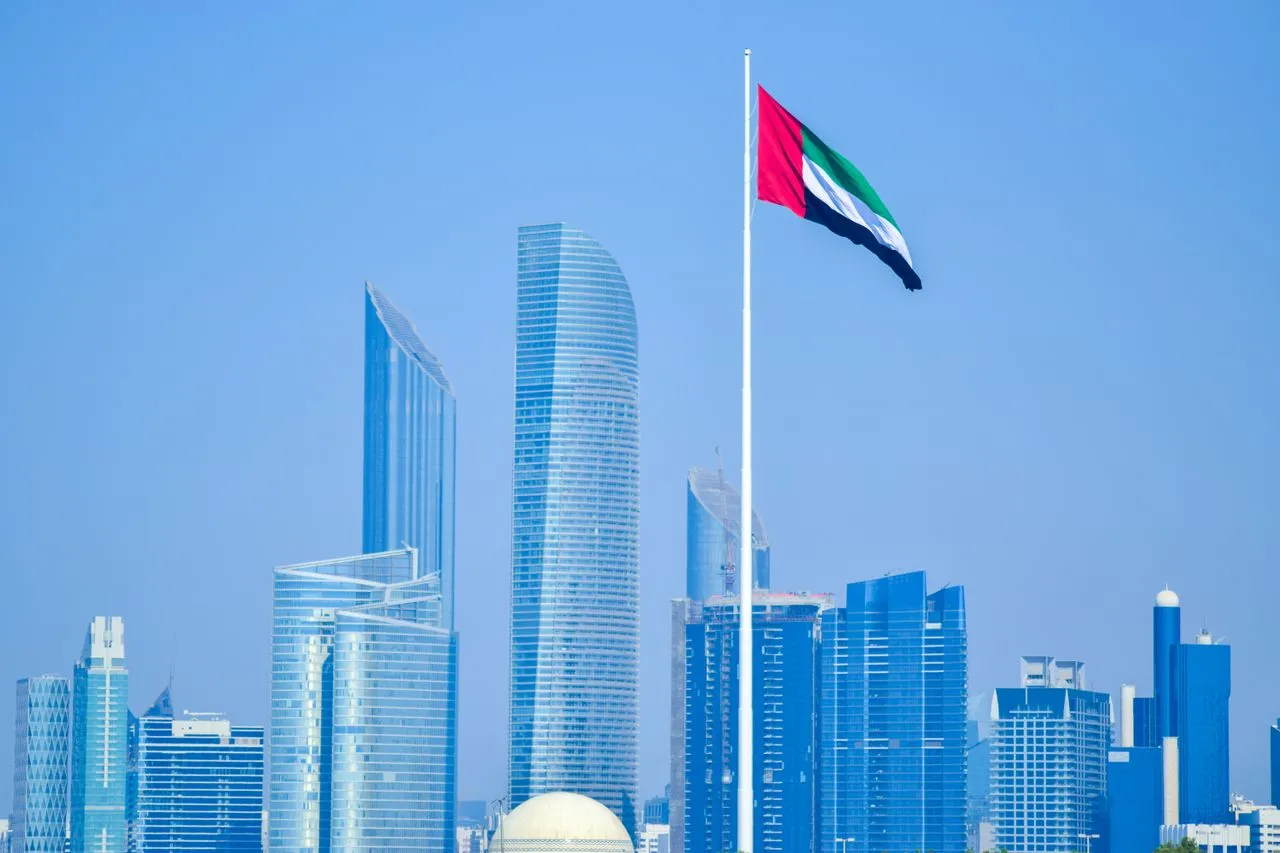 Skyline of modern skyscrapers with UAE flag prominently displayed against a clear blue sky.