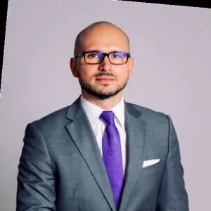 Lawyer in a formal suit with glasses, posing against a plain background.