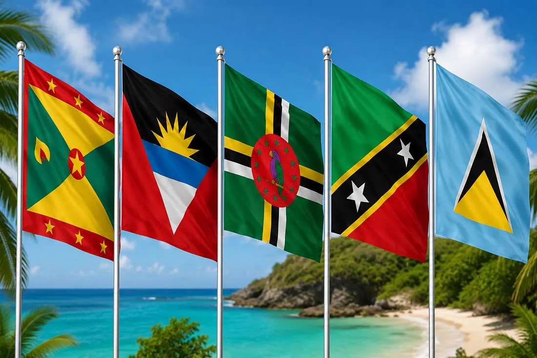Five national flags of Caribbean countries displayed on poles by a tropical beach with clear blue water in the background.