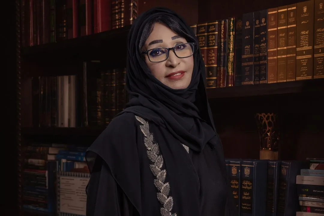 Legal professional posing in front of a bookshelf filled with legal texts, wearing a black abaya and glasses.