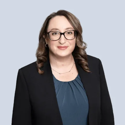 Attorney with shoulder-length hair wearing glasses and a black blazer, posing against a light gray background.